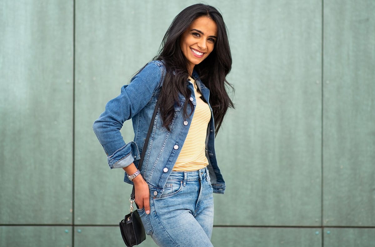 A woman with long, dark hair wearing a denim jacket and jeans. Her hands are on her hips and she is smiling,